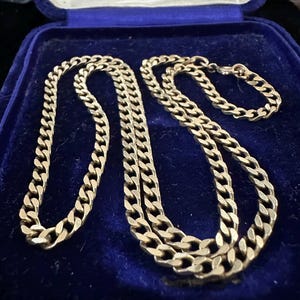 May include: A gold-colored chain necklace and bracelet set displayed inside a blue velvet jewelry box. The chain links are a curb style, and the set appears to be vintage or antique.
