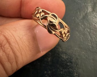 May include: A gold ring with a floral pattern. The ring has a repeating design of leaves and small flower clusters. The ring is held between fingers against a dark background.