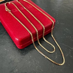 May include: A gold box chain necklace with a lobster clasp. The necklace is draped over a red jewellery box. The chain is made of small, rectangular links.