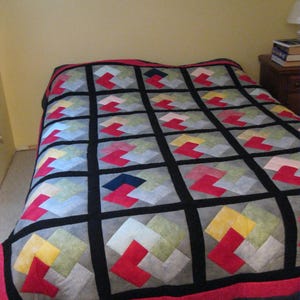Puede incluir: Una colcha con un patrón geométrico en tonos de rojo, amarillo, gris y azul. La colcha está compuesta por cuadrados con un borde negro.