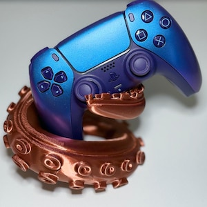 Könnte beinhalten: Ein metallisch blau-violetter Videospiel-Controller, der von einem kupferfarbenen Krakenarm-Ständer gehalten wird. Der Controller verfügt über Standardtasten, darunter ein Steuerkreuz, Analogsticks und das PlayStation-Logo in der Mitte.