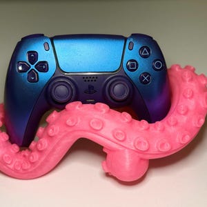May include: A metallic blue and purple video game controller resting inside a bright pink, textured octopus tentacle stand. The controller features standard directional buttons, two analog sticks, and the PlayStation logo in the centre.