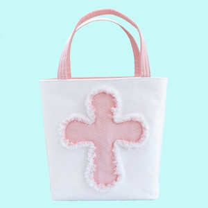 May include: White tote bag with pink cross applique and pink handles. The cross is outlined with a fluffy white trim. The bag is made of a sturdy fabric and is suitable for carrying small items.