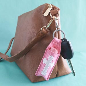 May include: A brown faux leather crossbody bag with a gold zipper and a pink polka dot fabric lip balm holder attached to the strap. The holder features a white lace cross applique and contains a light pink lip balm, alongside a black car key.