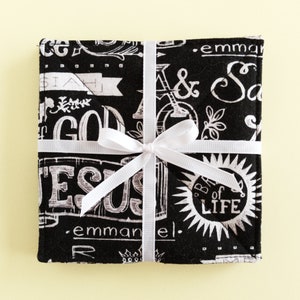 May include: A square stack of black fabric coasters featuring white printed text and illustrations including the words GOD, JESUS, and BREAD OF LIFE. The stack is tied together with a thin white ribbon tied in a bow at the center of the top.