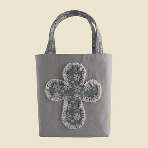 May include: A gray tote bag with a cross-shaped applique. The cross is made of a floral patterned fabric with a frayed edge. The handles are made of the same floral fabric. The bag is designed for carrying items.
