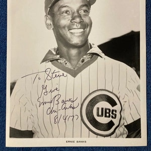 May include: Black and white photograph of Ernie Banks, a baseball player, wearing a Chicago Cubs uniform and cap. The image includes an autograph and the date 8/4/77. The Cubs logo is visible on the jersey.