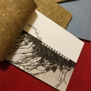 May include: A handmade book with a brown paper cover and the title "Notes to the Underground" printed in black. The book is open to reveal a black and white image of roots.