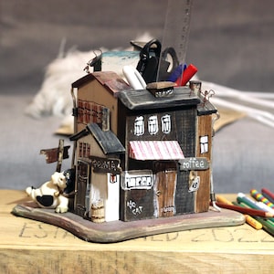 May include: A whimsical, hand-painted pen holder shaped like a multi-story building with a coffee shop. The holder features a red and white striped awning, a sign that says "coffee", and a small cow figurine. It holds pens, pencils, and a ruler.