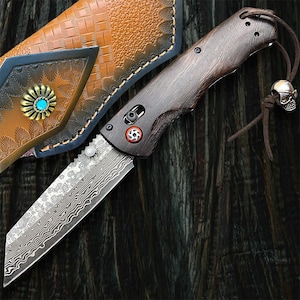 May include: A folding knife with a patterned blade and a dark wood handle. The knife features a decorative skull charm on a leather cord. A brown leather sheath with a turquoise accent is visible in the background.