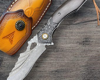 May include: A custom-made knife with a patterned blade and a dark wooden handle. The knife features ornate metalwork and is accompanied by a brown leather sheath with decorative accents. A leather cord is attached to the handle.