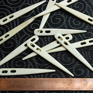 Puede incluir: Un grupo de 12 ganchillos de plástico blanco con una regla para la escala. Los ganchillos miden aproximadamente 10 cm de largo y tienen una punta puntiaguda. Los ganchillos tienen un agujero en la parte superior para enhebrar el hilo.