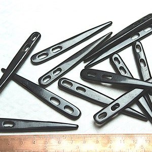 Puede incluir: Un conjunto de ballenas de cuello negras y puntiagudas, con agujeros ovalados, dispersas sobre una superficie blanca. Una regla proporciona la escala. Estos accesorios están diseñados para mantener los cuellos de las camisas.