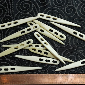 Puede incluir: Un grupo de 15 agujas de crochet de hueso blanco sobre un fondo negro. Las agujas están dispuestas de forma aleatoria y todas son del mismo tamaño. Una regla de metal se encuentra en la parte inferior de la imagen.