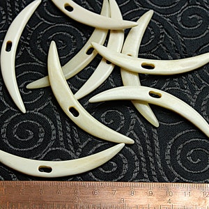 Puede incluir: Diez ganchos de ganchillo de hueso de color blanquecino, cada uno con un agujero en el centro, están esparcidos sobre una tela negra con un patrón de remolinos. Una regla es visible en la parte inferior de la imagen.