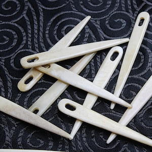 Puede incluir: Un grupo de diez agujas de hueso con agujeros para enhebrar. Las agujas están dispuestas en un patrón aleatorio sobre una tela negra con un diseño de espiral.