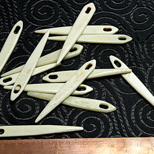 Puede incluir: Una colección de 15 agujas de hueso con agujeros para enhebrar. Las agujas son de diferentes longitudes y están dispuestas en un patrón aleatorio sobre un fondo de tela negra. Una regla es visible en la parte inferior de la imagen.