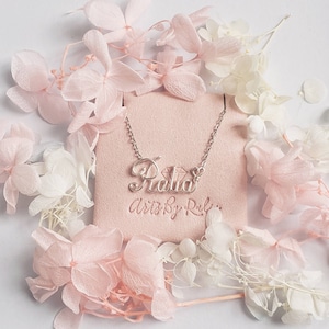 May include: A silver necklace with the name "Ralia" spelled out in cursive script. The necklace is displayed on a pink velvet background with white flowers.