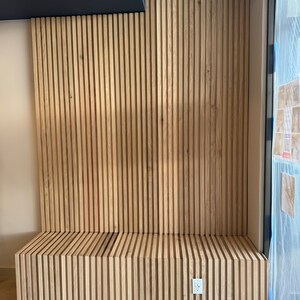 May include: A wooden bench with a slatted design, built into a wall with vertical wood slats. The bench is light brown and has a white electrical outlet on the wall.