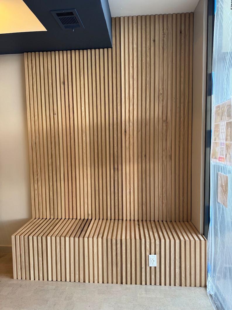 May include: A wooden bench with a slatted design, built into a wall with vertical wood slats. The bench is light brown and has a white electrical outlet on the wall.