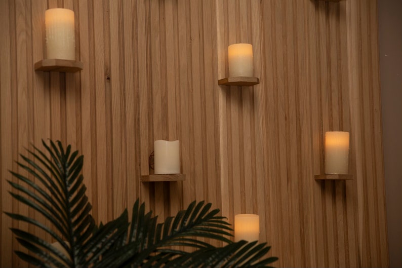 May include: A wooden wall with several small shelves holding lit candles. The shelves are mounted on the wall in a staggered pattern. A green plant is in the foreground.