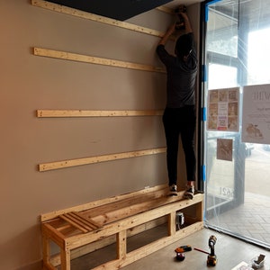 May include: A person is installing wooden shelves on a wall. A wooden bench with a slatted seat is in the foreground. The bench is unfinished and has a simple design. The person is using a power drill to attach the shelves to the wall.