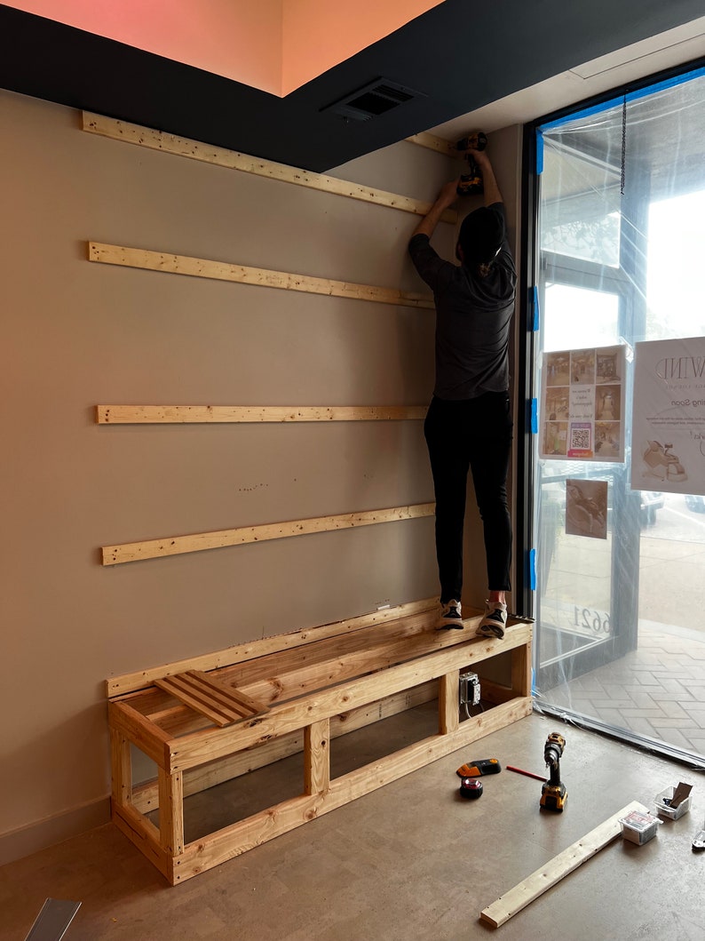 May include: A person is installing wooden shelves on a wall. A wooden bench with a slatted seat is in the foreground. The bench is unfinished and has a simple design. The person is using a power drill to attach the shelves to the wall.