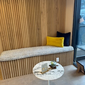 May include: A white marble round table with a white base sits on a light brown floor. A small glass globe with green plants sits on the table. A sign with the text "WTF" is on the table. A bench with a black and white patterned cushion sits against a wall with vertical wooden panels.