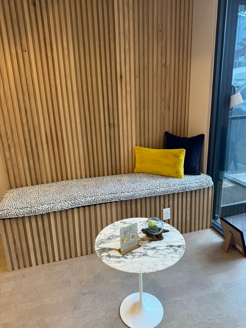May include: A white marble round table with a white base sits on a light brown floor. A small glass globe with green plants sits on the table. A sign with the text "WTF" is on the table. A bench with a black and white patterned cushion sits against a wall with vertical wooden panels.