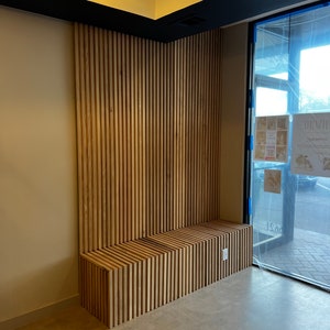 May include: A wooden bench with a slatted design is built into a wall with vertical wood slats. The wall is painted a light beige color and the bench is a natural wood color. The bench is in a room with a large window and a black ceiling trim.