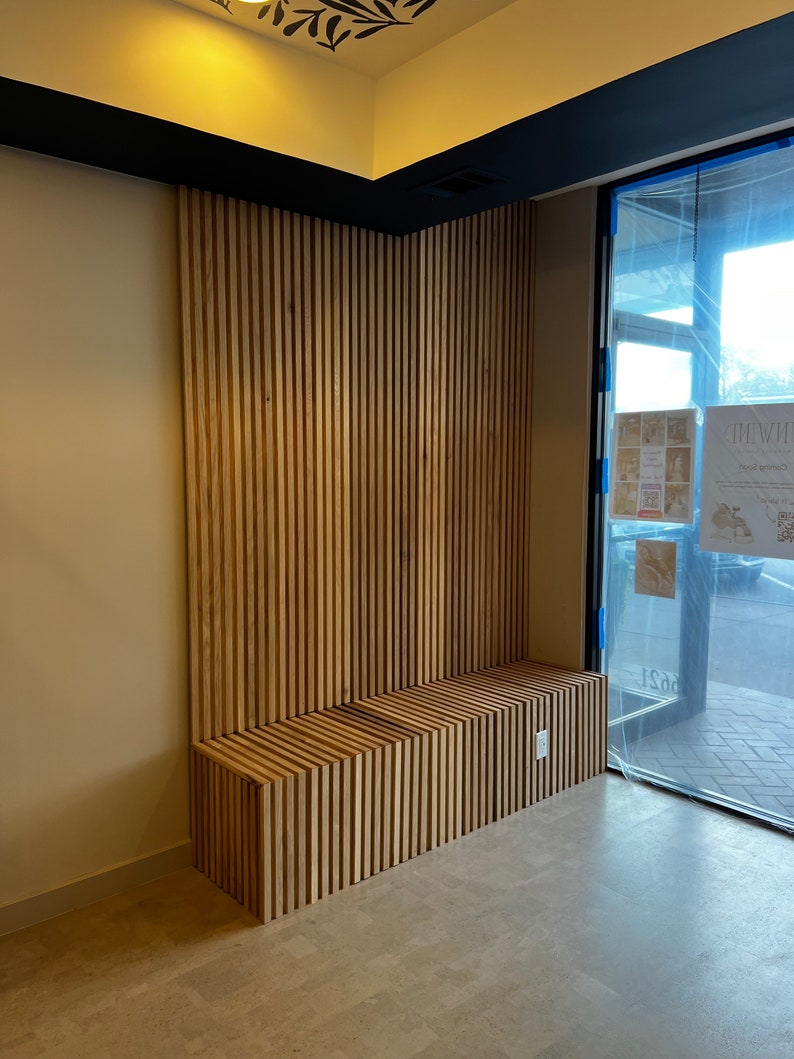 May include: A wooden bench with a slatted design is built into a wall with vertical wood slats. The wall is painted a light beige color and the bench is a natural wood color. The bench is in a room with a large window and a black ceiling trim.