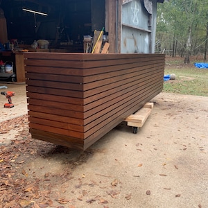 Peut inclure: Une longue jardinière rectangulaire en bois avec des lattes horizontales. La jardinière est en bois brun foncé et repose sur une plateforme en bois avec des roues.