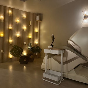 May include: A white massage chair with a gold accent and the word "Osaki" on the side. The chair is in a room with a wall of lit candles and a small white table with a bowl on top.