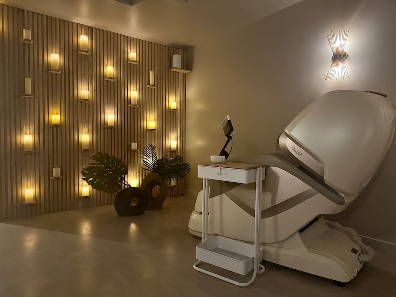 May include: A white massage chair with a gold accent and the word "Osaki" on the side. The chair is in a room with a wall of lit candles and a small white table with a bowl on top.