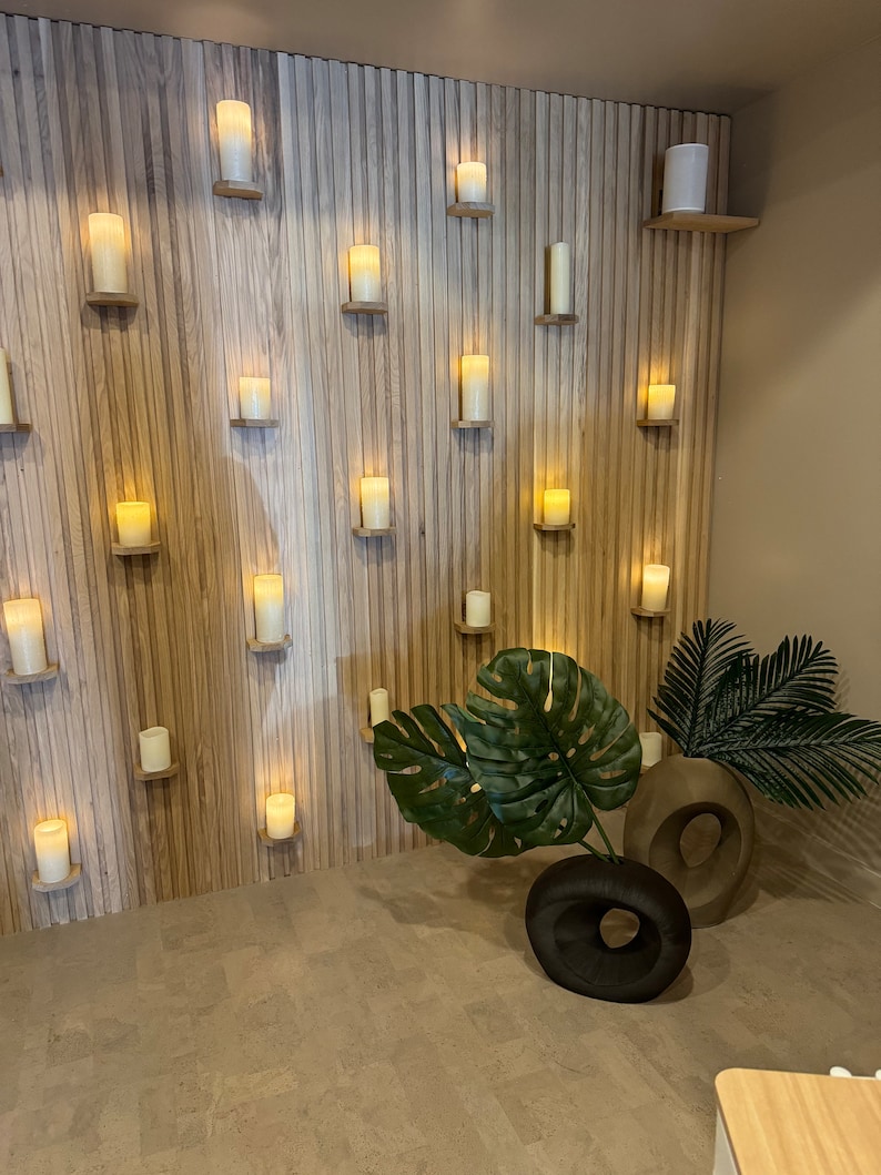 May include: A wall with wood paneling and a variety of white candles on shelves. There are two potted plants in the foreground, one with large green leaves and the other with smaller green leaves.