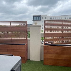 Peut inclure: Deux jardinières en bois avec des côtés à lattes et des écrans de confidentialité à lattes assortis derrière elles. Les jardinières sont sur un patio sur le toit avec de l'herbe artificielle.