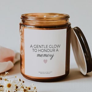 May include: An amber glass candle jar with a black lid and a white label featuring the text A GENTLE GLOW TO HONOUR A MEMORY, a small pink heart icon, and the text BREATHE IN THE LIGHT 8OZ SOY CANDLE. The candle is filled with soy wax.