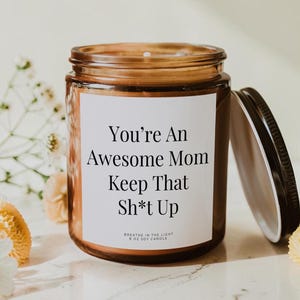 May include: A brown glass candle jar with a black lid and a white label featuring the black text: You're An Awesome Mom Keep That Sh*t Up. The label also includes the text BREATHE IN THE LIGHT 8 OZ SOY CANDLE at the bottom of the jar.