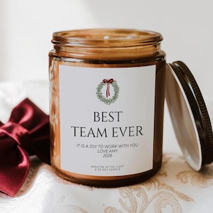 May include: An 8 oz soy candle in a brown glass jar with a black lid. The white label features a green wreath with a red bow, the text BEST TEAM EVER, IT IS A JOY TO WORK WITH YOU, LOVE AMY, 2026, and BREATHE IN THE LIGHT 8 OZ SOY CANDLE.