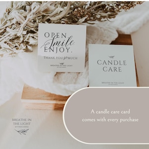 May include: Two white cards with black text. One card reads "OPEN. Smile. ENJOY. THANK YOU so much" and the other reads "CANDLE CARE". The cards are on a wooden surface with dried foliage. The brand name "BREATHE IN THE LIGHT" is also visible.