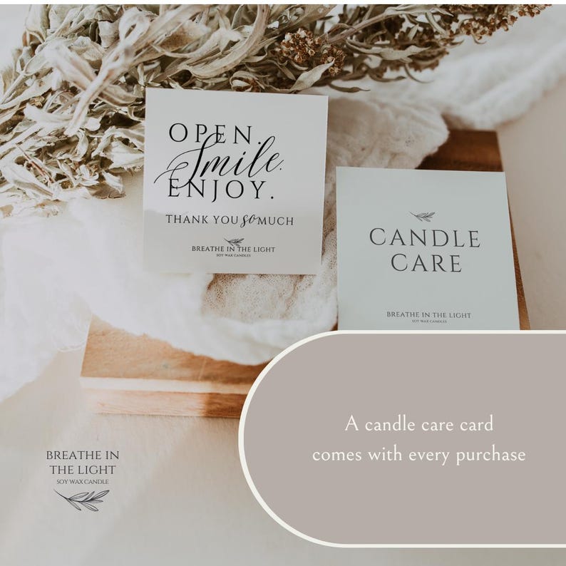 May include: Two white cards with black text. One card reads "OPEN. Smile. ENJOY. THANK YOU so much" and the other reads "CANDLE CARE". The cards are on a wooden surface with dried foliage. The brand name "BREATHE IN THE LIGHT" is also visible.