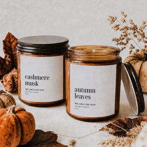 May include: Two amber glass soy wax candles with black lids. One label reads Cashmere Musk, Breathe in the Light, 8 oz. The other label reads Autumn Leaves, Breathe in the Light, 8 oz. The candles are surrounded by dried autumn leaves and small pumpkins.
