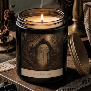 May include: A lit candle in a black glass jar with a gold-toned metal lid. The label features an illustration of a stone archway in a forest with a ghostly figure and the text "beyond the veil" printed in a simple, lowercase serif font.