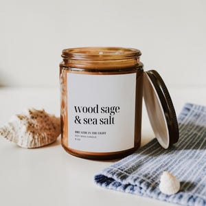 May include: A brown glass jar candle with a black lid, labeled "wood sage & sea salt." The candle sits next to a seashell and a blue and white striped cloth. The candle is 8 oz.