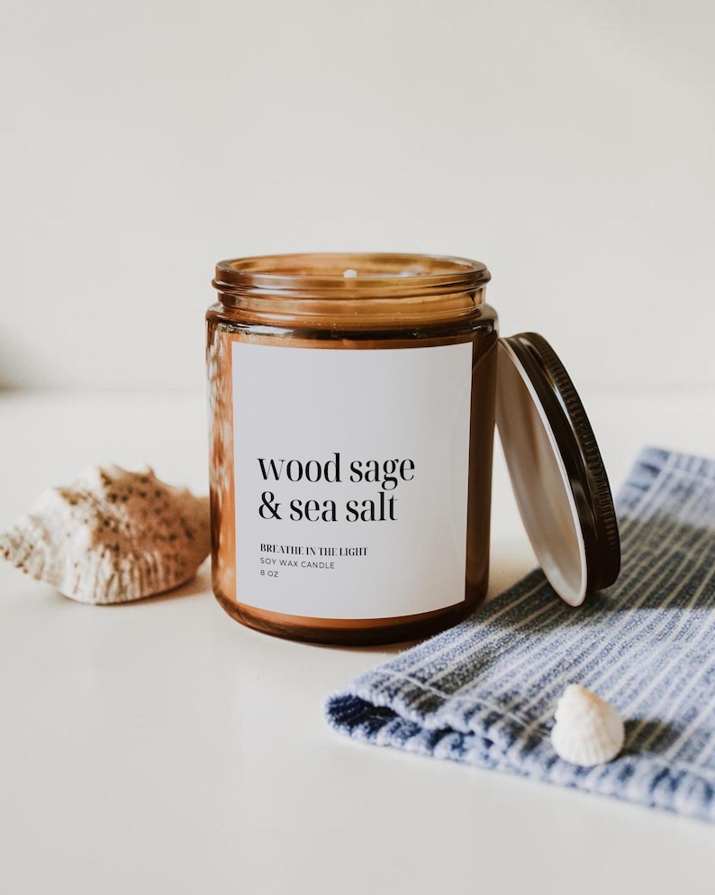 May include: A brown glass jar candle with a black lid, labeled "wood sage & sea salt." The candle sits next to a seashell and a blue and white striped cloth. The candle is 8 oz.