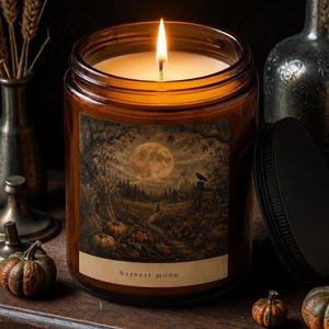 Peut inclure: Une bougie allumée dans un pot en verre ambré cylindrique avec un couvercle en métal noir. L'étiquette présente une illustration détaillée d'une pleine lune au-dessus d'un champ de citrouilles, d'une forêt et d'un corbeau, avec le texte harvest moon.