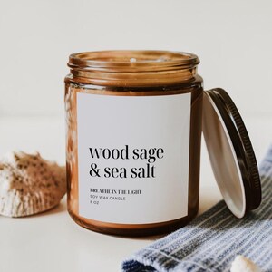 May include: A soy wax candle in a clear glass jar with a black lid. The label reads "wood sage & sea salt" and "Breathe in the light". The candle is 6 oz. A seashell and a blue and white striped cloth are in the background.