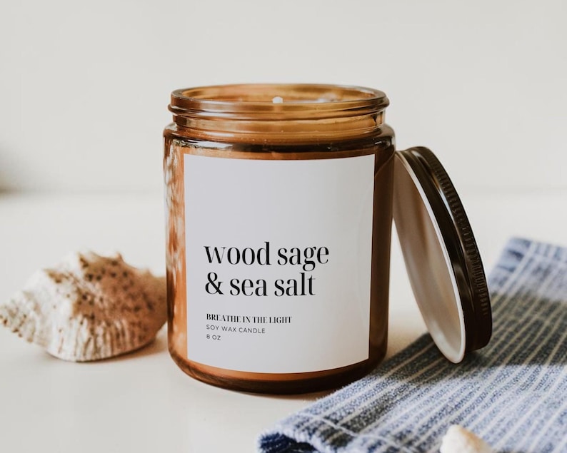 May include: A soy wax candle in a clear glass jar with a black lid. The label reads "wood sage & sea salt" and "Breathe in the light". The candle is 6 oz. A seashell and a blue and white striped cloth are in the background.