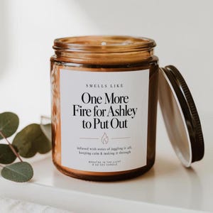 May include: An 8 oz soy candle in a brown glass jar with a black lid. The white label features the text: SMELLS LIKE One More Fire for Ashley to Put Out, infused with notes of juggling it all, keeping calm & making it through, BREATHE IN THE LIGHT.