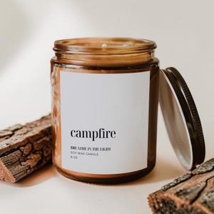 May include: A brown glass jar soy wax candle with a white label featuring the text campfire, BREATHE IN THE LIGHT, SOY WAX CANDLE, and 8 oz. The candle has a matching brown metal lid placed to the right side of the jar.